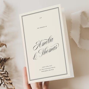 May include: A cream wedding invitation with black text, held by a hand. The text reads "Amelia & Thomas" in elegant script. A sprig of dried foliage and a white ribbon are also in the image.