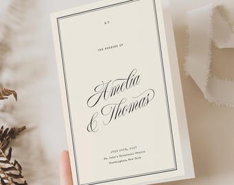 Könnte beinhalten: Eine cremefarbene Hochzeitseinladungskarte mit schwarzem Text, der Amelia & Thomas, July 25th, 2017, St. Jude's Episcopal Church, Huntington, New York lautet, eingefasst in einen dünnen schwarzen rechteckigen Rahmen, begleitet von einem getrockneten Farn und weißem Band.