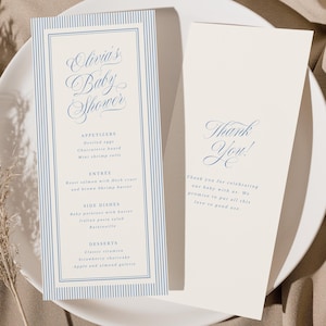 May include: Baby shower menu and thank you card on a white plate. The menu features a blue striped border and elegant script that reads "Olivia's Baby Shower." The thank you card has the words "Thank You!" in a similar script. Beige fabric is in the background.