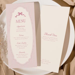 May include: A baby shower menu and thank you card on a white plate. The menu features a pink bow and vertical stripes, listing appetizers, entrees, and desserts. The thank you card has a script font and the text "Thank You".