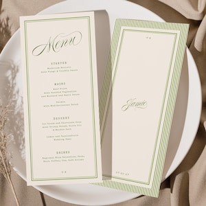 Peut inclure: Deux cartons de menu crème avec des bordures vertes et du texte, l'un intitulé "Menu" et l'autre "Jamie", reposent sur une assiette blanche. Le fond est un tissu beige avec une plante séchée.