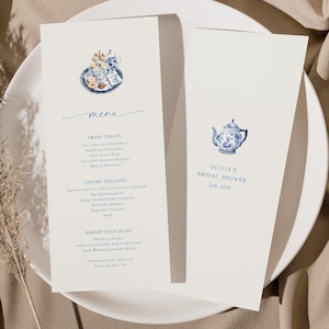 Op de afbeelding: Twee crèmekleurige menukaarten op een wit bord. Eén kaart toont een aquarel illustratie van een theeservies en het woord "menu". De andere kaart zegt "Olivia's Bridal Shower" en "Juni 2024".
