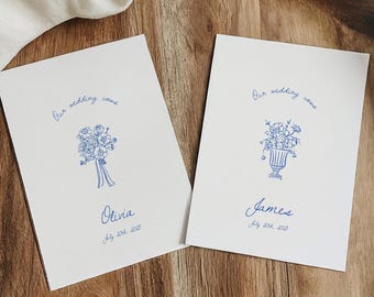 Könnte beinhalten: Zwei weiße Karten mit blauer Tinte. Jede Karte hat eine Blumenillustration und den Text "Our wedding name". Eine Karte sagt "Olivia" und die andere sagt "James".