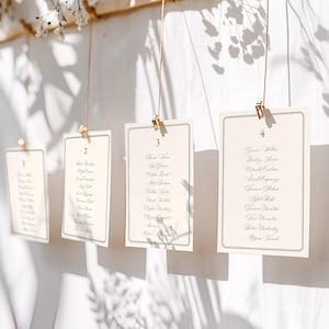 Può includere: Biglietti color crema per il tableau de mariage con bordo verde, sospesi con clip dorate. Ogni biglietto è numerato e presenta un elenco di nomi in elegante calligrafia. I biglietti sono appesi su uno sfondo bianco, con accenti floreali.