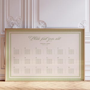 May include: A framed seating chart with the text "Please find your seat" in elegant script. The chart has a light pink background with rows of names and table assignments, framed in gold and green. The setting is a room with white paneling and a wooden floor.