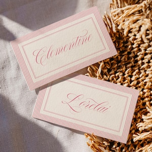 Puede incluir: Dos tarjetas de mesa rectangulares con los nombres "Clementine" y "Lorelai" en elegante escritura cursiva. Las tarjetas tienen un fondo crema con un borde a rayas rosa y un marco interior rosa. Sobre una superficie tejida.