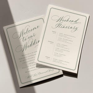 May include: Two off-white rectangular cards with dark green borders and elegant script text. One card reads Welcome to our Wedding, the other Weekend Itinerary, detailing events from Friday to Sunday with specific times and locations for each.