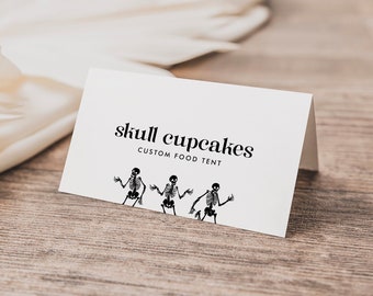 Könnte beinhalten: Eine weiße Karte mit schwarzem Text, der "skull cupcakes" und "custom food tent" lautet. Am unteren Rand der Karte befinden sich drei schwarze Skelettfiguren.
