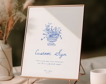 May include: A gold-framed sign with a blue floral illustration and the text "Custom Sign". The sign is on a gold stand and sits on a beige table.