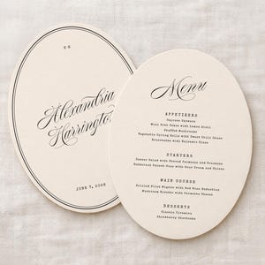 May include: Two off-white, oval-shaped wedding menu cards. One card features the name "Alexandria Harrington" in elegant script. The other card lists a menu with appetizers, starters, main course, and desserts. The text is black.