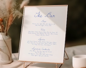May include: A gold-framed sign with a white background and blue text that reads "The Bar". The sign lists various beer, wine, spirits, and signature cocktails. The sign is on a gold stand and is sitting on a table with a white vase and a white cup.