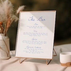 Puede incluir: Un cartel con marco dorado, fondo blanco y texto azul que dice "The Bar". El cartel enumera varias cervezas, vinos, licores y cócteles de autor. El cartel está en un soporte dorado y está sobre una mesa con un jarrón blanco y una taza blanca.