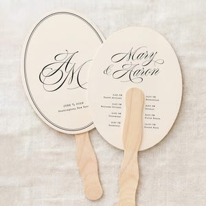 May include: Two ivory-colored wedding fans with wooden handles. One fan features the initials "J M" in elegant script, the other displays "Mary & Aaron" with a wedding timeline. The fans are oval-shaped with a thin black border.