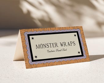 Könnte beinhalten: Eine gefaltete Tischkarte aus Papier mit einem beigen Rand mit schwarzen vertikalen Linien, einem hellvioletten Wellenrahmen und einem cremefarbenen Mittelfeld. Der Text lautet MONSTER WRAPS und Custom Food Tent, ergänzt durch vier kleine schwarze Sterne.