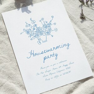 Puede incluir: Una tarjeta de invitaci&oacute;n blanca con ilustraciones florales azules y texto. La tarjeta dice "Housewarming party" en escritura cursiva, con detalles del evento e informaci&oacute;n de contacto. El dise&ntilde;o presenta un jarr&oacute;n de flores.