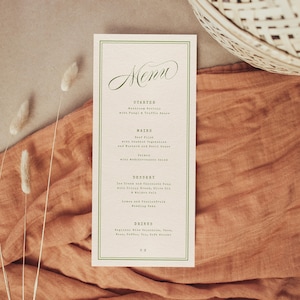 May include: A cream-coloured menu card with a green border and elegant script font. The menu lists starters, mains, dessert, and drinks. The card is set on a rust-coloured fabric with decorative dried plants and a woven basket.