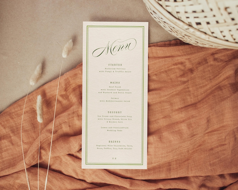 May include: A cream-coloured menu card with a green border and elegant script font. The menu lists starters, mains, dessert, and drinks. The card is set on a rust-coloured fabric with decorative dried plants and a woven basket.