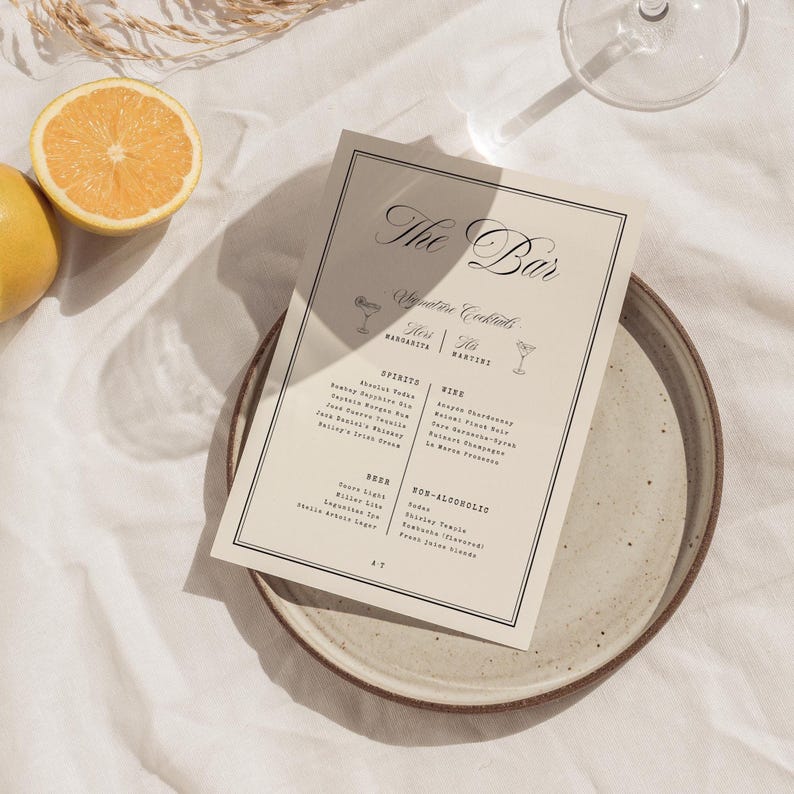 May include: A rectangular bar menu card with black text on a cream background, featuring the title The Bar, Signature Cocktails, and sections for Spirits, Wine, Beer, and Non-Alcoholic drinks, framed by a thin black border on a ceramic plate.