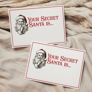 May include: Two white cards with red borders and a black and white illustration of Santa Claus with his finger to his lips. The text on the cards reads "Your Secret Santa is..."