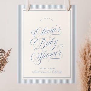 May include: A baby shower sign with a cream background and blue pinstripe border. The sign reads "Welcome to Olivia's Baby Shower" in elegant script, with the date and location below. The sign is suspended by white ribbons.