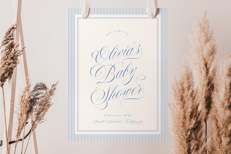 May include: A baby shower sign with a cream background and blue pinstripe border. The sign reads "Welcome to Olivia's Baby Shower" in elegant script, with the date and location below. The sign is suspended by white ribbons.