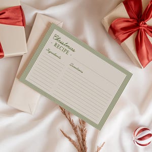 May include: A cream-colored recipe card with green striped borders and the text "Christmas RECIPE" is displayed. Two wrapped gifts with red satin bows and a decorative ornament are also present, all on a white satin surface.