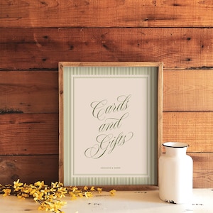 May include: A rectangular wooden frame containing a sign with a cream background, green vertical stripes, and green cursive text that reads Cards and Gifts, with the smaller text Caroline & Henry printed at the bottom of the sign.