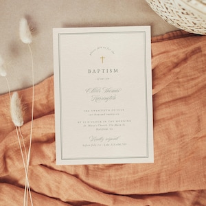 May include: A cream-colored baptism invitation with a gold cross and elegant script. The invitation includes the word "BAPTISM" and other details. The card is set on a peach-colored fabric with dried flowers.