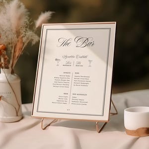 May include: A rectangular bar menu sign in a thin rose gold frame, featuring the text The Bar, Signature Cocktails, Spirits, Wine, Beer, and Non-Alcoholic. The sign is displayed on a wire stand next to a white vase with dried flowers and a small white cup.