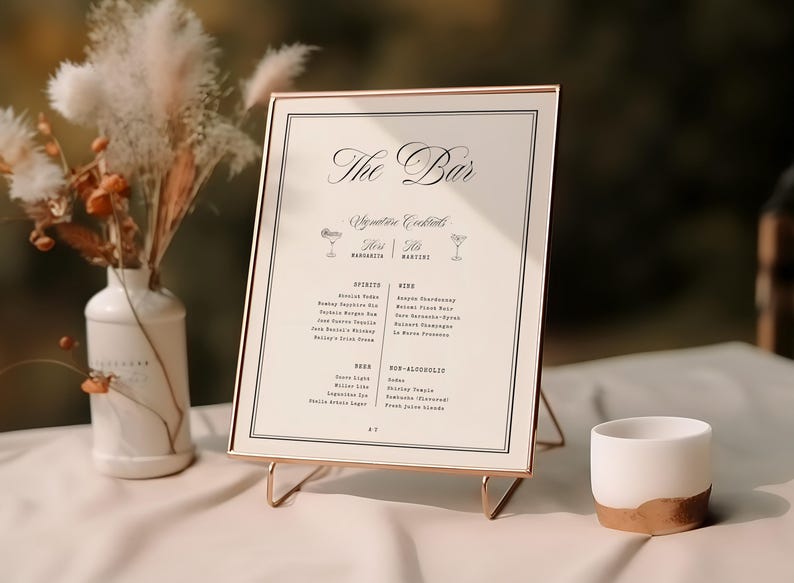 May include: A rectangular bar menu sign in a thin rose gold frame, featuring the text The Bar, Signature Cocktails, Spirits, Wine, Beer, and Non-Alcoholic. The sign is displayed on a wire stand next to a white vase with dried flowers and a small white cup.