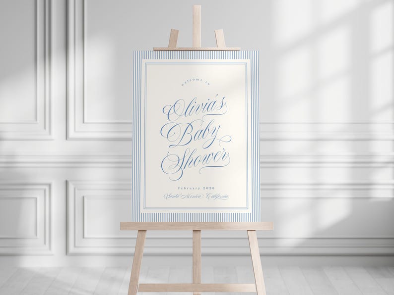 May include: A baby shower sign on a wooden easel. The sign has a cream-colored background with blue pinstripe borders and text that reads "Olivia's Baby Shower" in elegant script. The sign also includes the date and location.