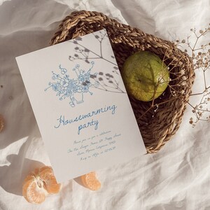 Puede incluir: Una tarjeta de invitaci&oacute;n blanca con ilustraciones florales azules y las palabras "Housewarming party". La tarjeta est&aacute; colocada en una cesta de mimbre con una fruta verde y flores secas. Rodajas de naranja est&aacute;n esparcidas.
