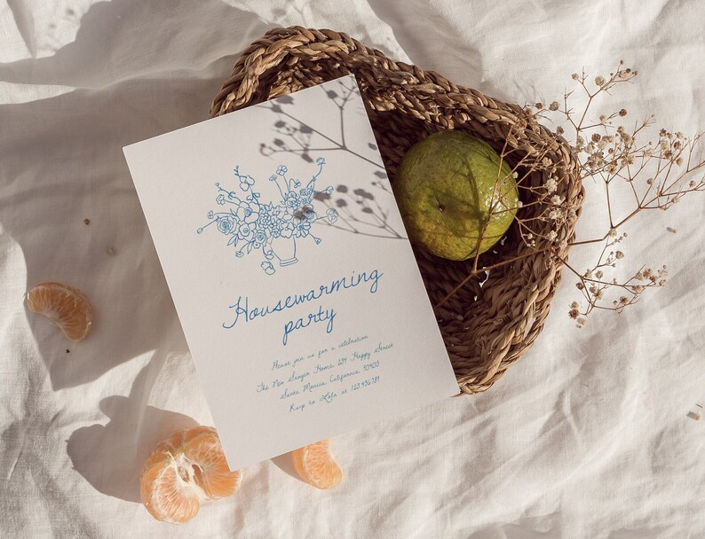 Puede incluir: Una tarjeta de invitaci&oacute;n blanca con ilustraciones florales azules y las palabras "Housewarming party". La tarjeta est&aacute; colocada en una cesta de mimbre con una fruta verde y flores secas. Rodajas de naranja est&aacute;n esparcidas.