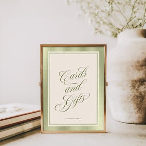 May include: A rectangular sign in a thin metallic frame featuring a cream-colored background with a green striped border. The text "Cards and Gifts" is written in a dark green cursive script, with "CAROLINE & HENRY" printed in small block letters below.