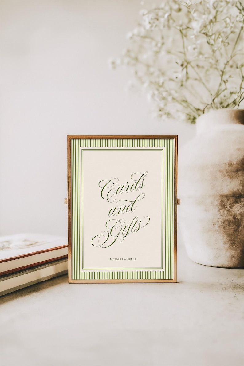 May include: A rectangular sign in a thin metallic frame featuring a cream-colored background with a green striped border. The text "Cards and Gifts" is written in a dark green cursive script, with "CAROLINE & HENRY" printed in small block letters below.