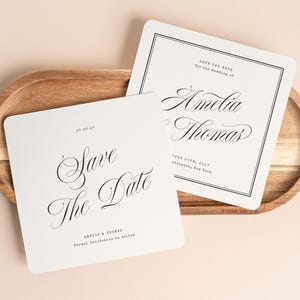 Puede incluir: Dos tarjetas cuadradas blancas de "Save the Date" con texto negro y un borde decorativo sobre una bandeja de madera. Las tarjetas presentan una escritura elegante, incluyendo nombres y una fecha de boda. La bandeja es ovalada.