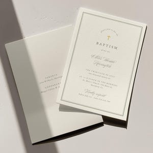 May include: Two off-white baptism invitations with elegant gold text and a cross symbol. One invitation is open, revealing the event details, while the other is closed. The invitations are on a light-colored surface.