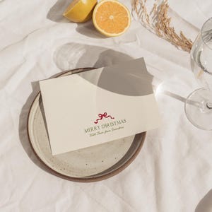 May include: A cream-colored Christmas card with a red bow design and the text "MERRY CHRISTMAS With Love from Grandma" sits on a speckled ceramic plate. The plate is on a white cloth, with a halved orange and a glass of water in the background.