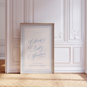 May include: A framed baby shower sign with a gold border and blue text. The sign reads "welcome to Olivia's Baby Shower" with the date February 2020 and the location, Walnut Creek, California. The sign is displayed in a room with white paneling and a wooden floor.