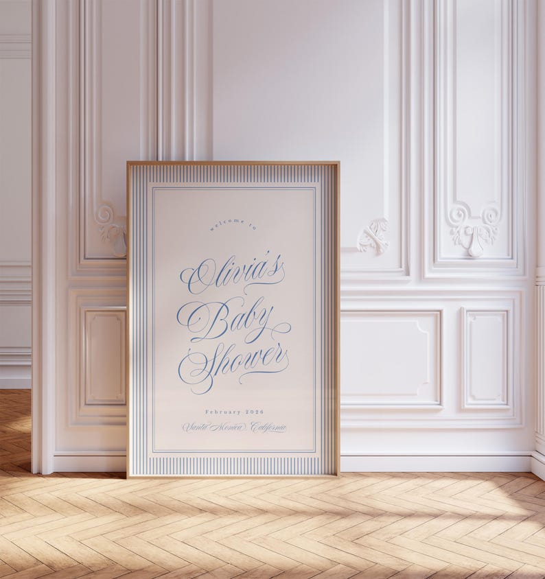 May include: A framed baby shower sign with a gold border and blue text. The sign reads "welcome to Olivia's Baby Shower" with the date February 2020 and the location, Walnut Creek, California. The sign is displayed in a room with white paneling and a wooden floor.