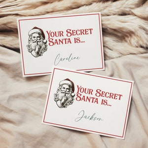 May include: Two white cards with red borders and a Santa Claus illustration. The text on the cards reads "Your Secret Santa is..." followed by a name. The first card says "Caroline" and the second card says "Jackson".