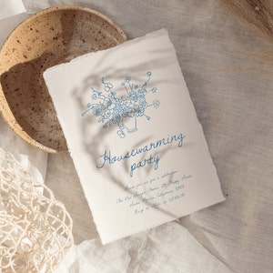 Puede incluir: Una invitaci&oacute;n color crema con una ilustraci&oacute;n floral azul y las palabras "Housewarming party". La invitaci&oacute;n est&aacute; en papel texturizado, con un borde rasgado. Un cuenco de cer&aacute;mica y una bolsa de red est&aacute;n en el fondo.