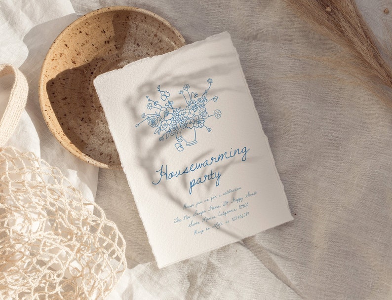 Puede incluir: Una invitaci&oacute;n color crema con una ilustraci&oacute;n floral azul y las palabras "Housewarming party". La invitaci&oacute;n est&aacute; en papel texturizado, con un borde rasgado. Un cuenco de cer&aacute;mica y una bolsa de red est&aacute;n en el fondo.