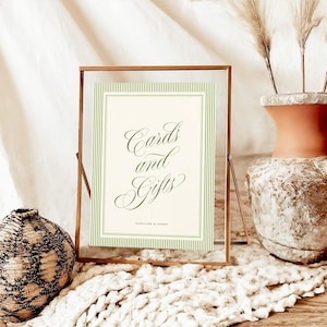 May include: A rectangular glass frame with a thin copper border containing a cream-colored sign with green pinstriped borders. The sign features the dark green cursive text "Cards and Gifts" above the smaller text "Jocelyn & David".