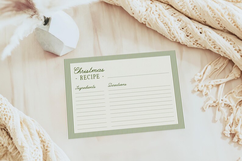 May include: A cream-colored recipe card with green striped borders and the words "Christmas Recipe" in elegant script. The card has spaces for ingredients and directions, perfect for holiday baking. A cozy, cream-colored knitted blanket and decorative vase are in the background.
