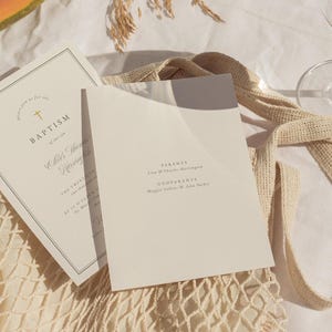 May include: Two off-white baptism invitations with gold text and a cross graphic. One invitation is open, revealing the text "Parents" and "Godparents." The other invitation is closed, with the word "Baptism" visible.