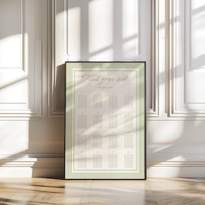 May include: A framed seating chart with the text "Find your seat" in elegant script. The chart has a cream-colored background with a green striped border and a black frame. The chart is leaning against a white wall.