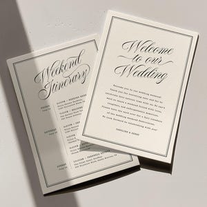 Può includere: Due cartoncini rettangolari color bianco sporco con testo nero e bordi a doppia linea. Un cartoncino riporta la scritta Welcome to our Wedding in carattere corsivo, l'altro Weekend Itinerary. Il testo include dettagli dell'evento e i nomi.