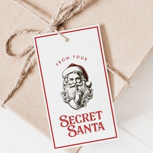May include: White gift tag with a red border. The tag features a black and white illustration of Santa Claus with his finger to his lips. The text "From your Secret Santa" is printed in red.
