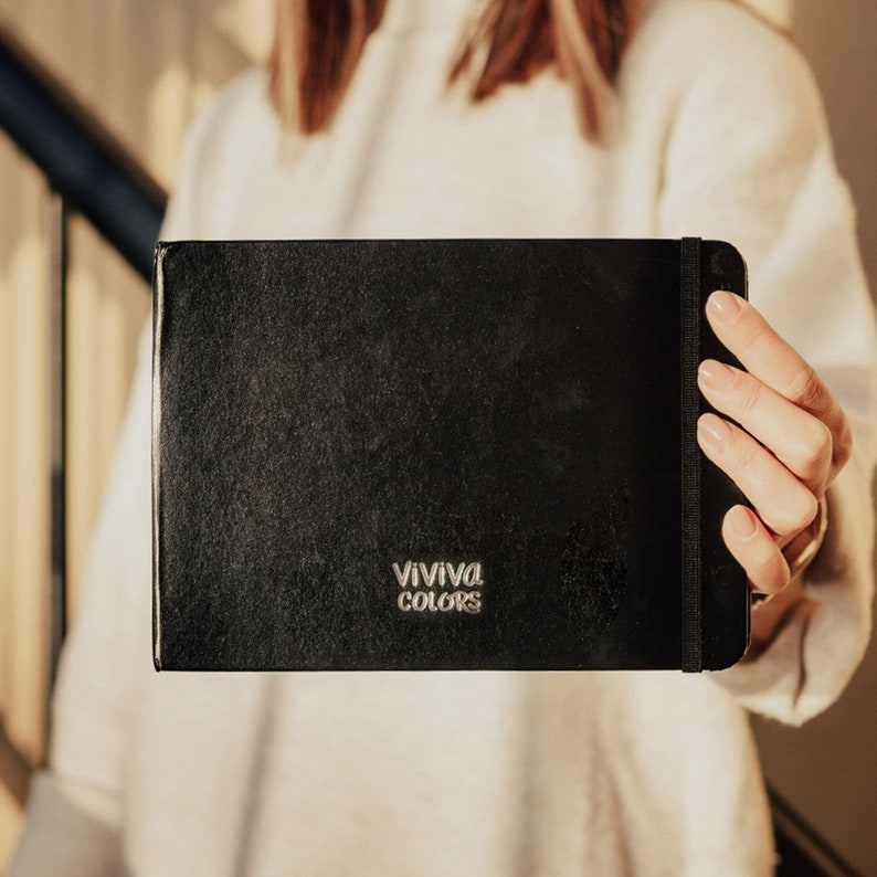 Peut inclure: Un carnet de croquis noir avec un logo argent&eacute; en relief qui dit "VIVIVA COLORS".